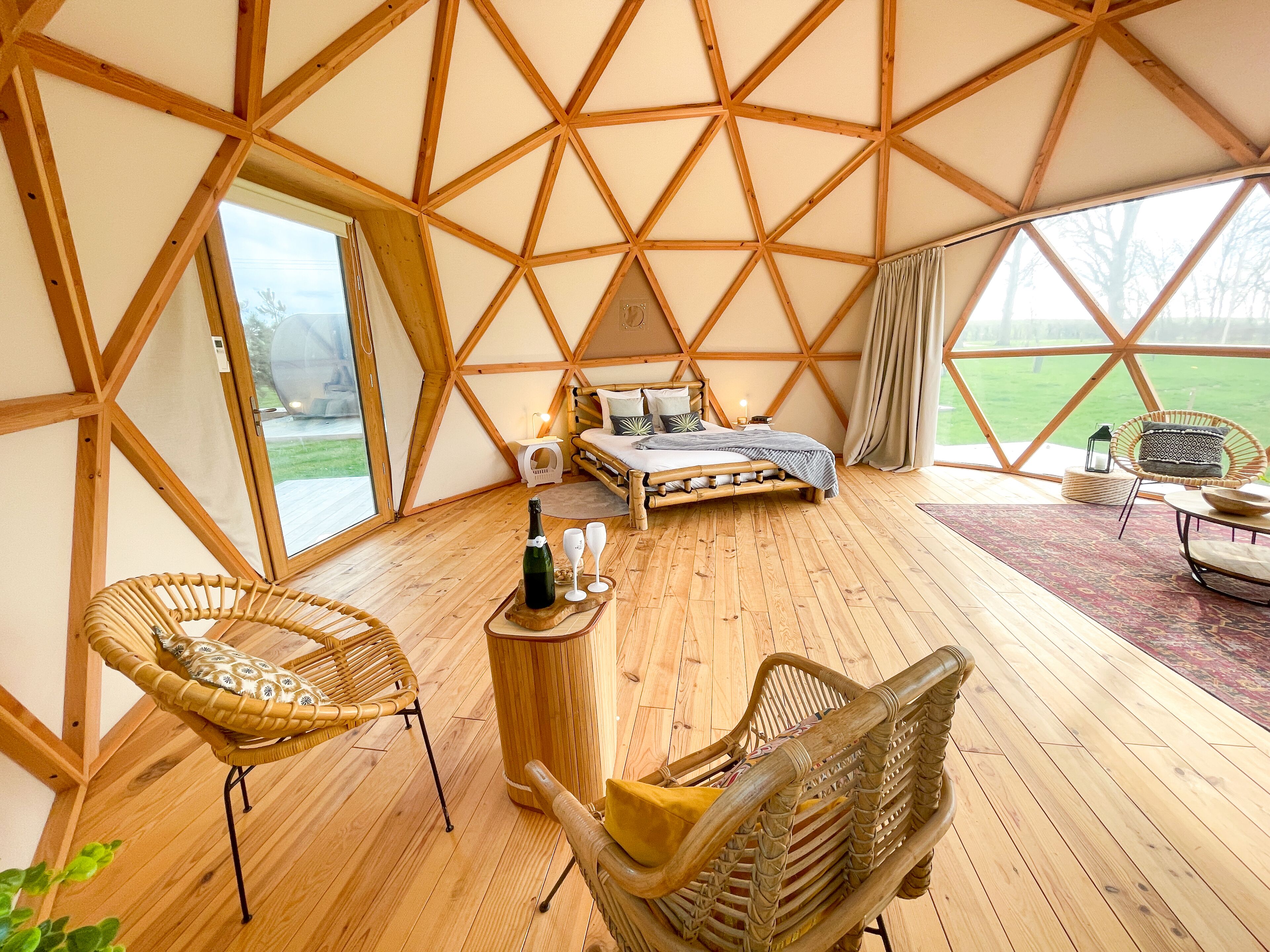 A spacious tent with a wooden floor, a bed, a wicker chair, and a table with a bottle and glasses.