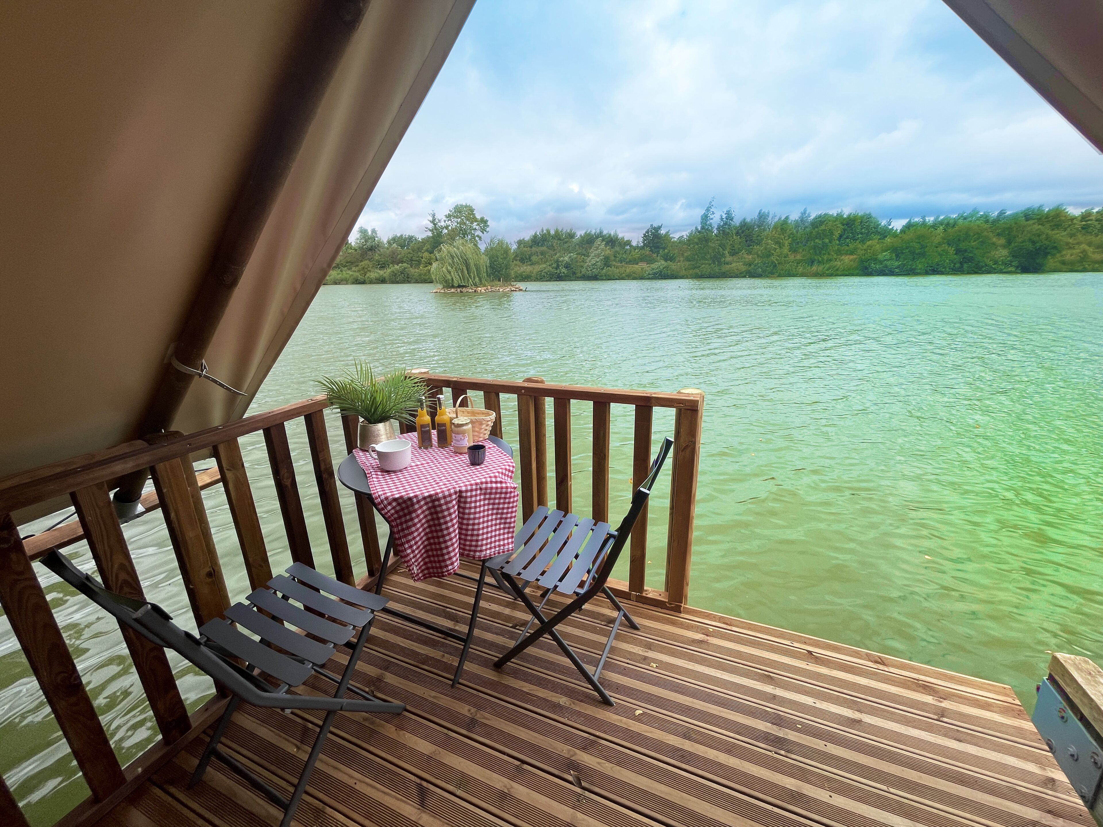 A wooden deck with a table and chairs, overlooking a body of water with a forested shoreline.