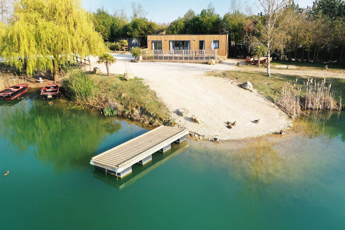 Un muelle de madera que se adentra en una masa de agua tranquila, un pequeño edificio con porche y un sauce llorón cerca.