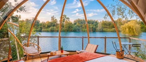 Acogedora habitación con cama, una mesa y sillas, con vista a un lago rodeado de árboles y cielo despejado.