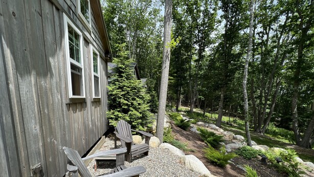 Una cabaña de madera con dos sillas Adirondack afuera, rodeada de un área boscosa con un sendero de grava y rocas.