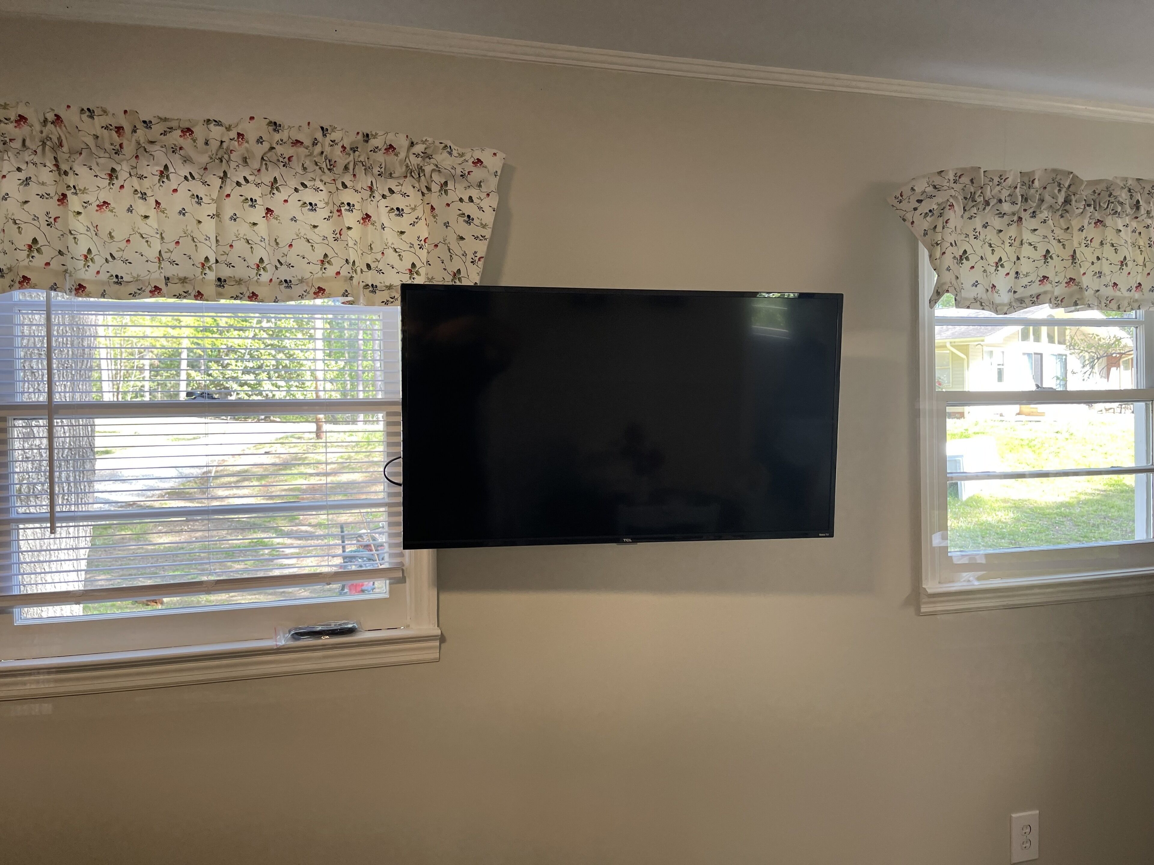 Un televisor montado en la pared entre dos ventanas con cortinas florales.