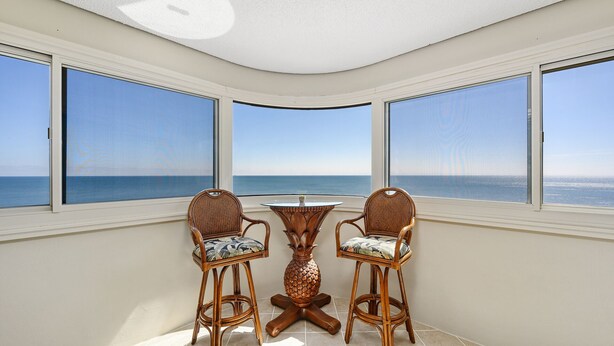 Ein Eckzimmer mit zwei Korbstühlen und einem kleinen Tisch, große Fenster mit Blick aufs Meer und eine weiße Decke.