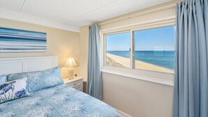 Ein Schlafzimmer mit Bett, Nachttisch, Lampe, Fenster mit Meerblick und einem Bild an der Wand.