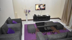 Uma sala de estar com sofás cinza, uma mesa de centro preta, uma televisão de tela plana e um vaso com flores secas.