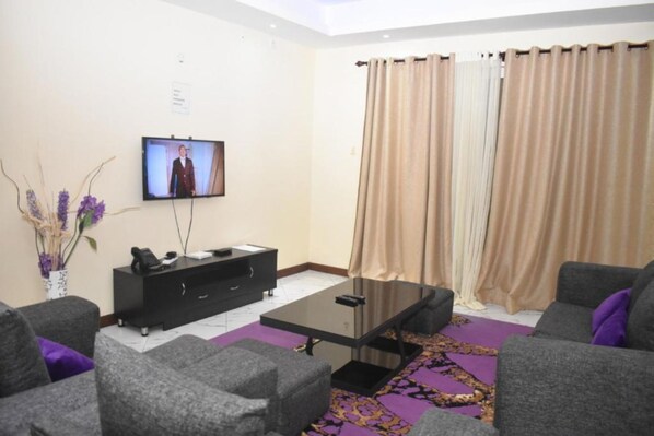 A modern living room with grey sofas, a black coffee table, a flat-screen TV mounted on the wall, and a decorative vase with purple flowers.