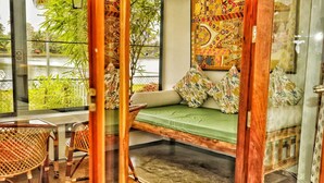 Une pièce avec un banc rembourré de couleur verte, des chaises en osier, une décoration murale à motifs et une vue sur la verdure à l’extérieur.