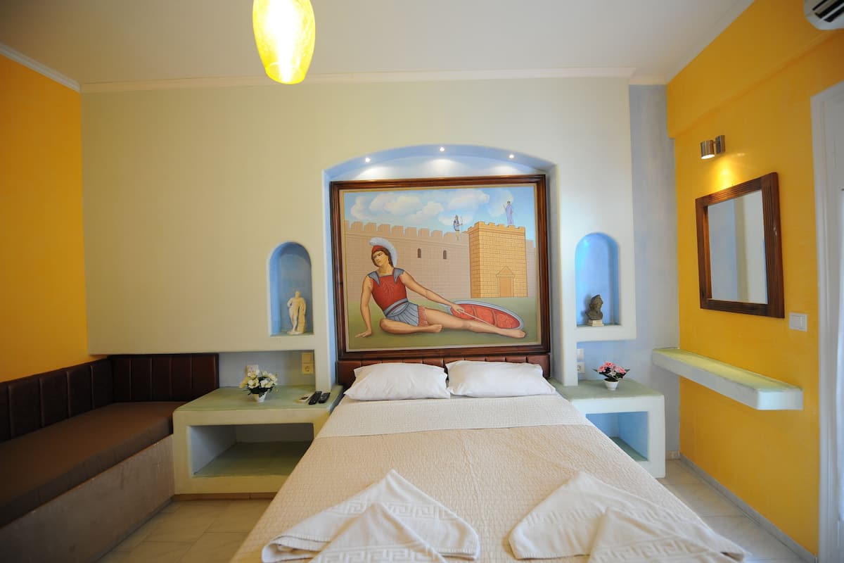 Habitación de hotel con cama, banca, mesita de noche, un cuadro, espejo y una pequeña mesa con un florero.
