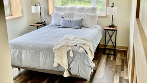 A bedroom with a large bed, two bedside tables with lamps, a window with a view of greenery, and wooden flooring. - Private Ridge top Cabin/House (Avoca)