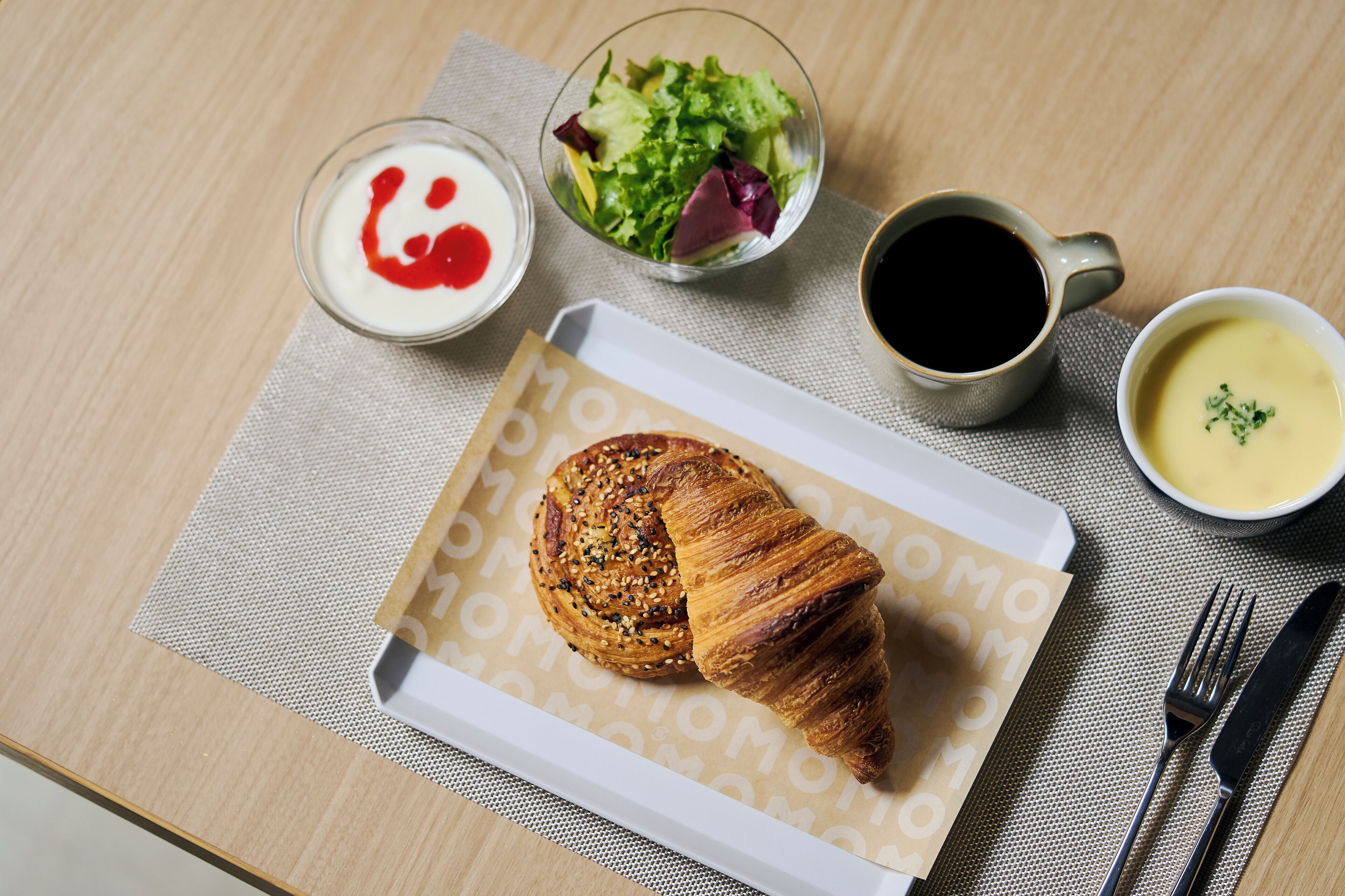 A croissant on a paper towel, a bowl of soup, a cup of coffee, a glass of milk with a red syrup, and a salad on a table.