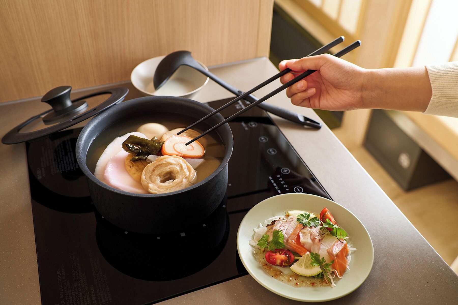 Una cocina con una olla de comida en la estufa, un plato de pescado y verduras, y una mano sosteniendo palillos.