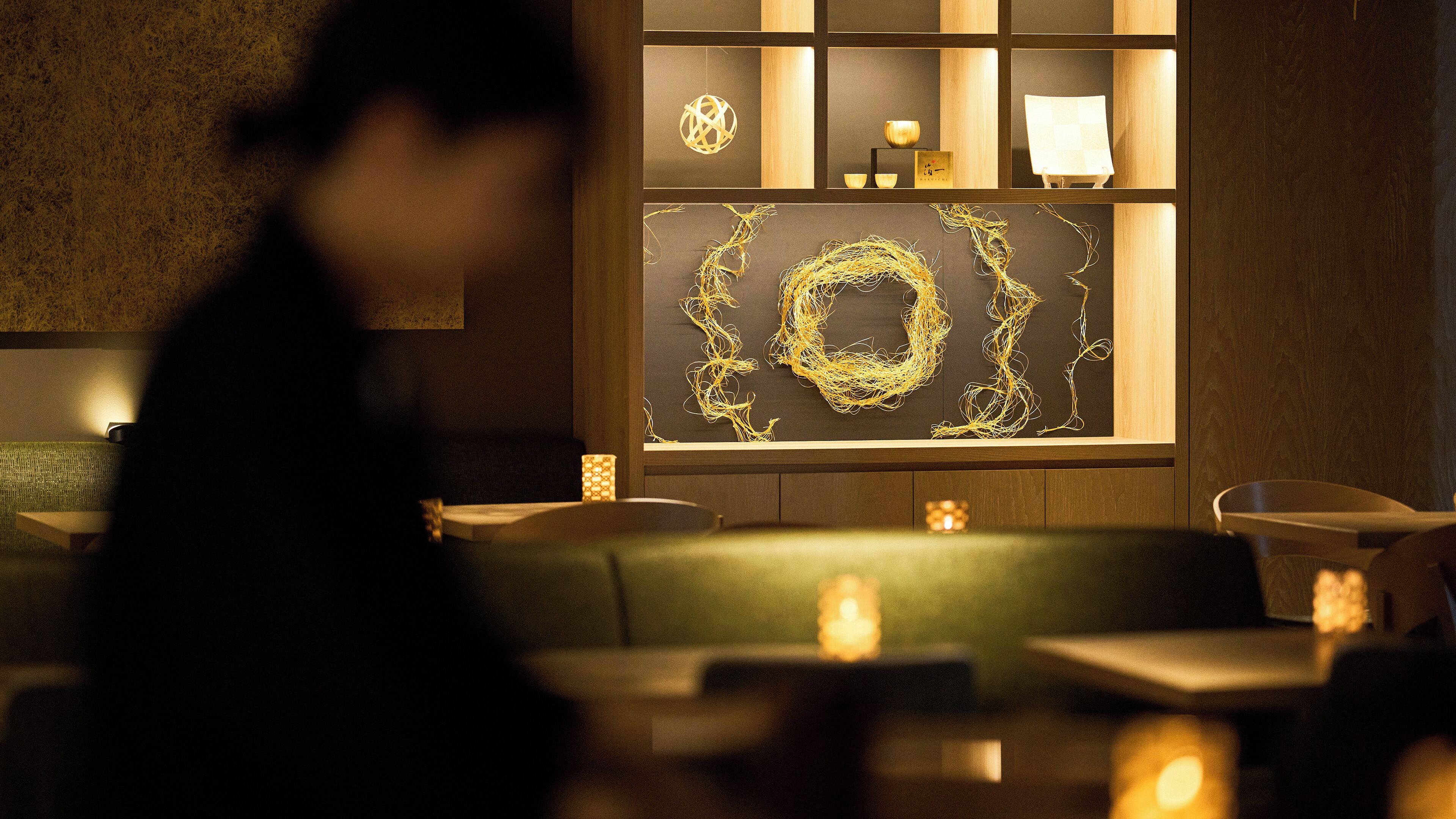 A dimly lit interior with a green sofa and tables, a decorative wall with a golden circular design, and a wooden shelf with various items.