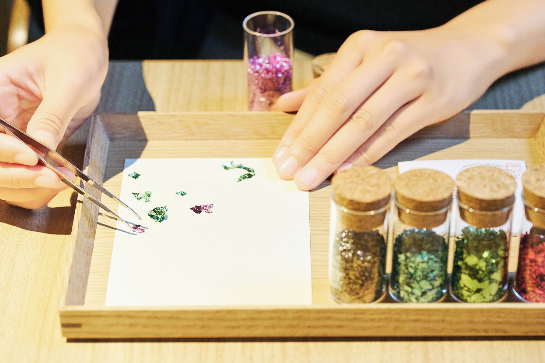 A person using tweezers to place small green and purple objects on a white surface, with jars of various colored powders nearby.
