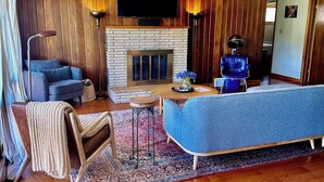 A living room with a fireplace, blue sofas, wooden flooring, and a ceiling fan. - Relax & Recharge: Mid-Century Modern Home with Sauna in Spring Green (Spring Green)