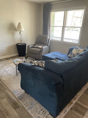 Un salon avec un canapé bleu, un fauteuil, une petite table avec des magazines, une lampe et une fenêtre avec des stores.