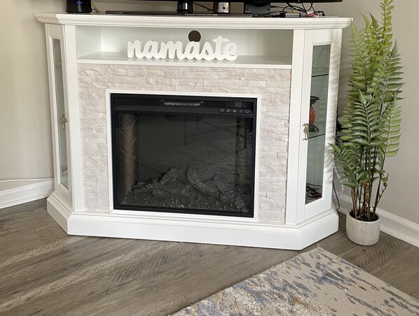 A white fireplace with a glass door, a decorative sign above it, and a potted plant to the right.