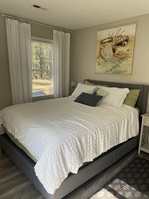 Un dormitorio con una cama grande, una ventana con cortinas, una mesita de noche y un cuadro colgado en la pared.