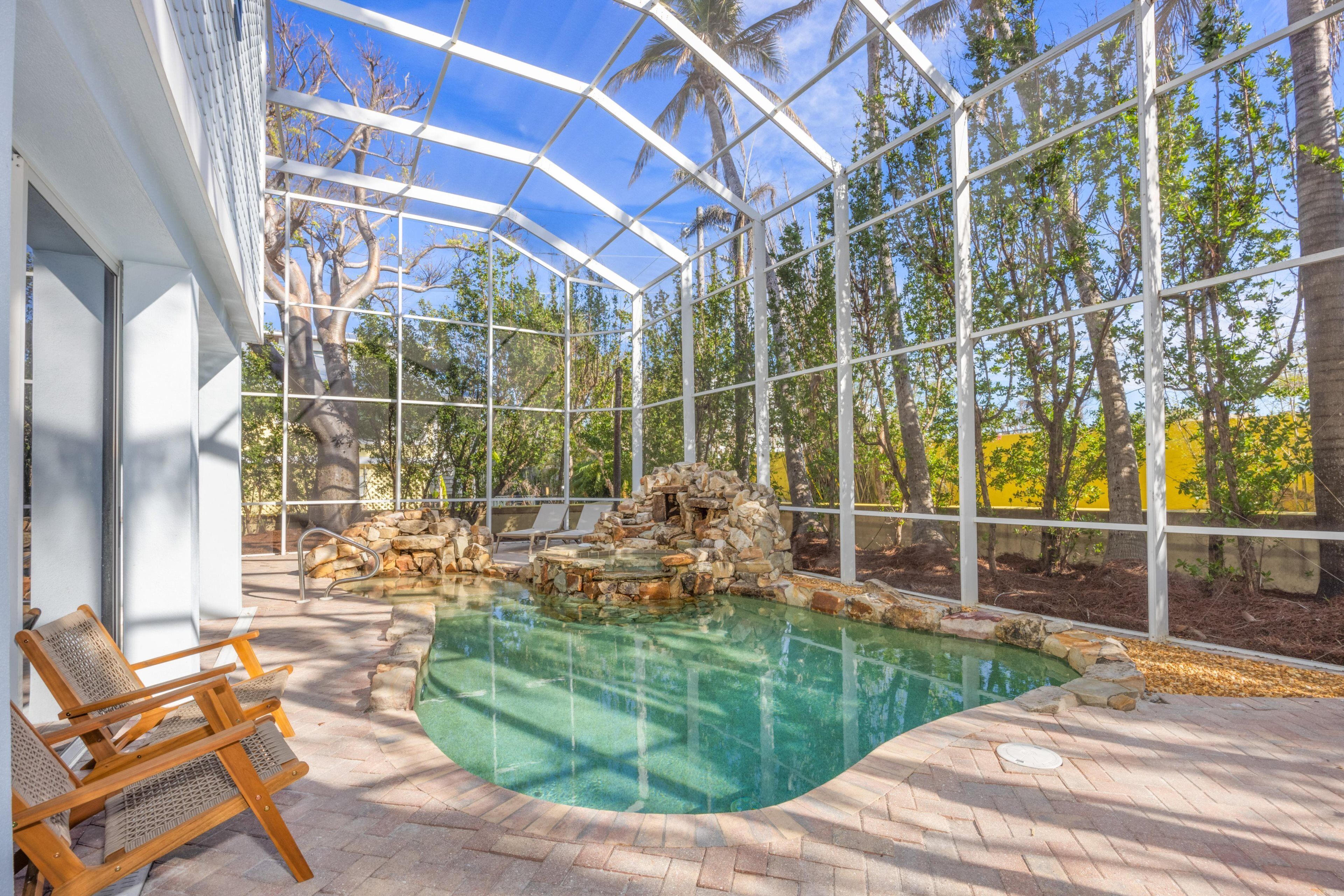 A glass-enclosed swimming pool with stone features, wooden lounge chairs, and a view of trees and sky.