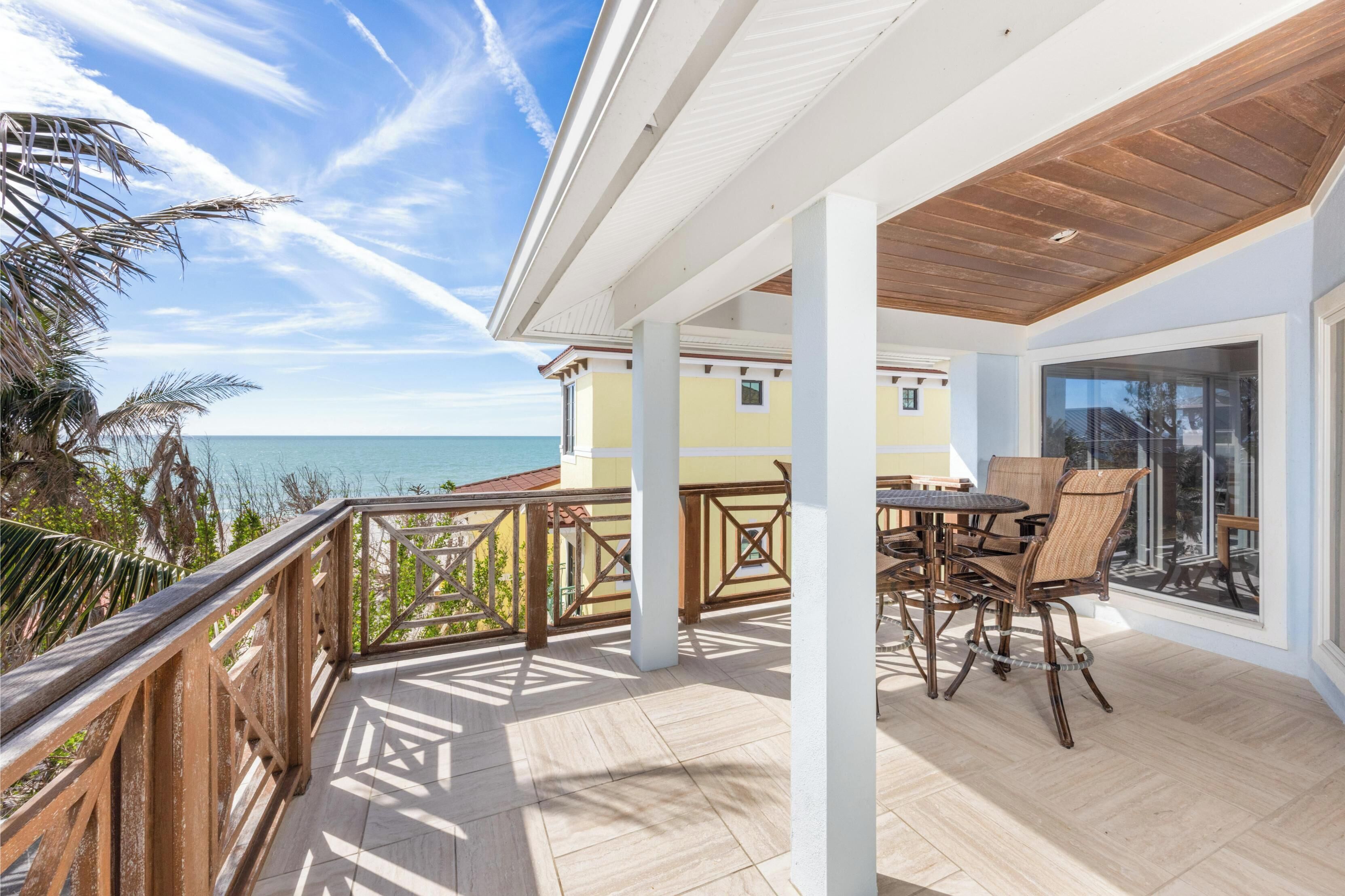A covered outdoor area with a wooden table and chairs, a view of the ocean, and a building in the background.