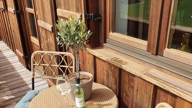 Une terrasse en bois avec une table et des chaises rondes en osier, une petite plante en pot et une fenêtre donnant sur l’extérieur.