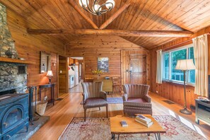 Interior acolhedor de uma cabana com lareira, paredes de madeira, área de jantar e uma janela grande.
