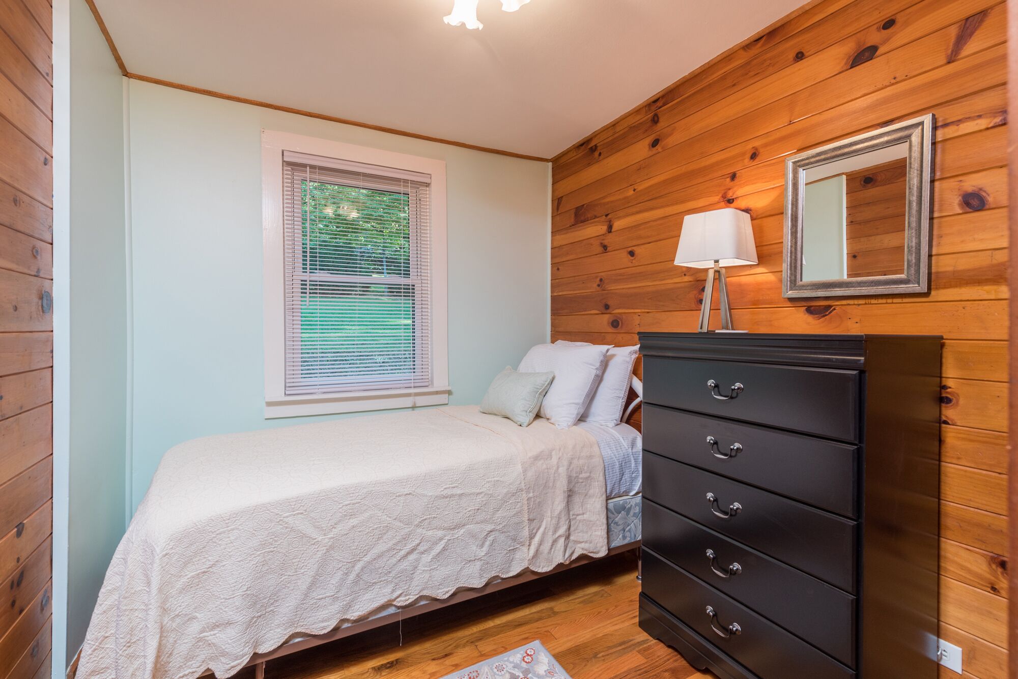 A bedroom with a bed, a dresser, a window with blinds, and a mirror on the wall.