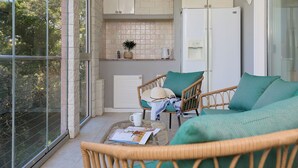 A modern kitchen with white cabinets, a built-in oven, and a coffee machine. There are wicker chairs with green cushions and a glass table with a magazine and a white mug. - Hedera Estate, Hedera A3 (Dubrovnik)