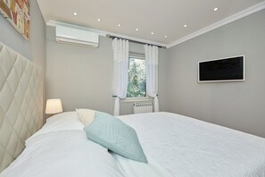 A bedroom with a bed, a bedside lamp, a window with curtains, an air conditioning unit, a radiator, and a TV mounted on the wall. - Hedera Estate, Hedera A3 (Dubrovnik)
