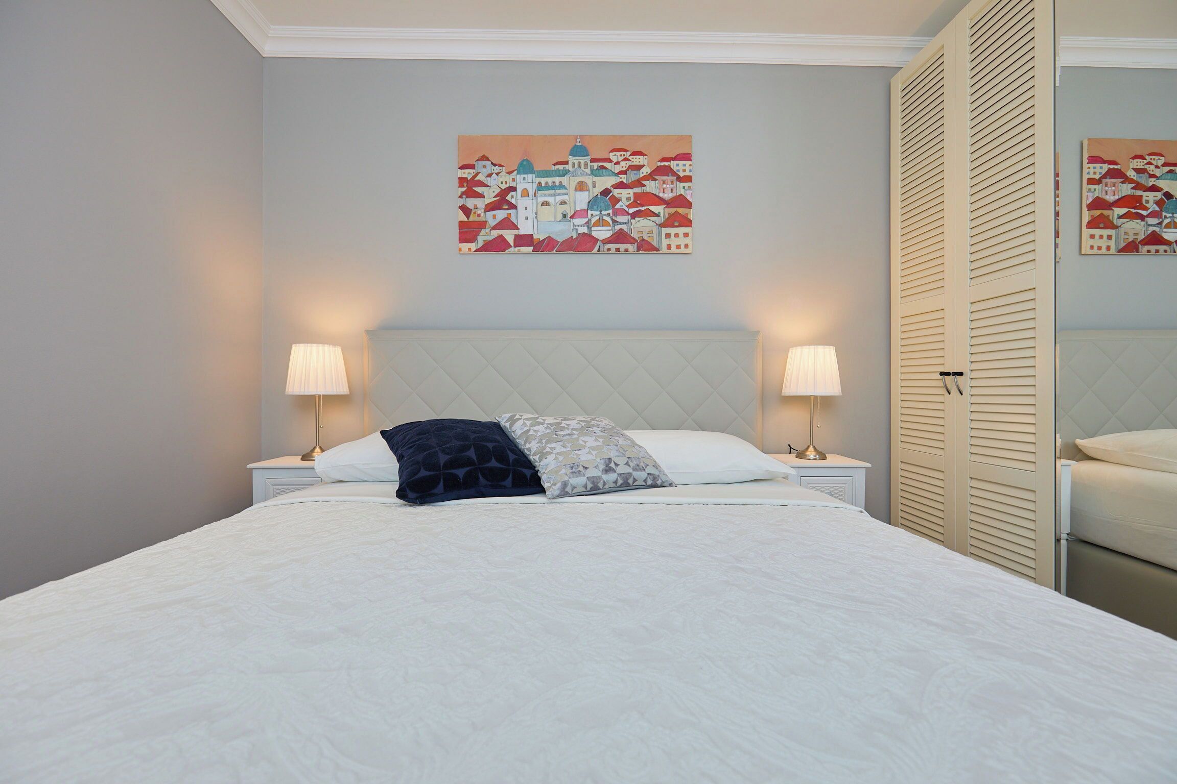 A neatly made bed with a white duvet and two pillows, flanked by bedside tables with lamps, in a room with a light blue wall and a cityscape painting above the bed.