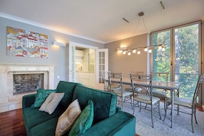 A living room with a green sofa, a fireplace, a dining area with chairs and a table, and a large window with a view of trees. - Hedera Estate, Hedera A3 (Dubrovnik)