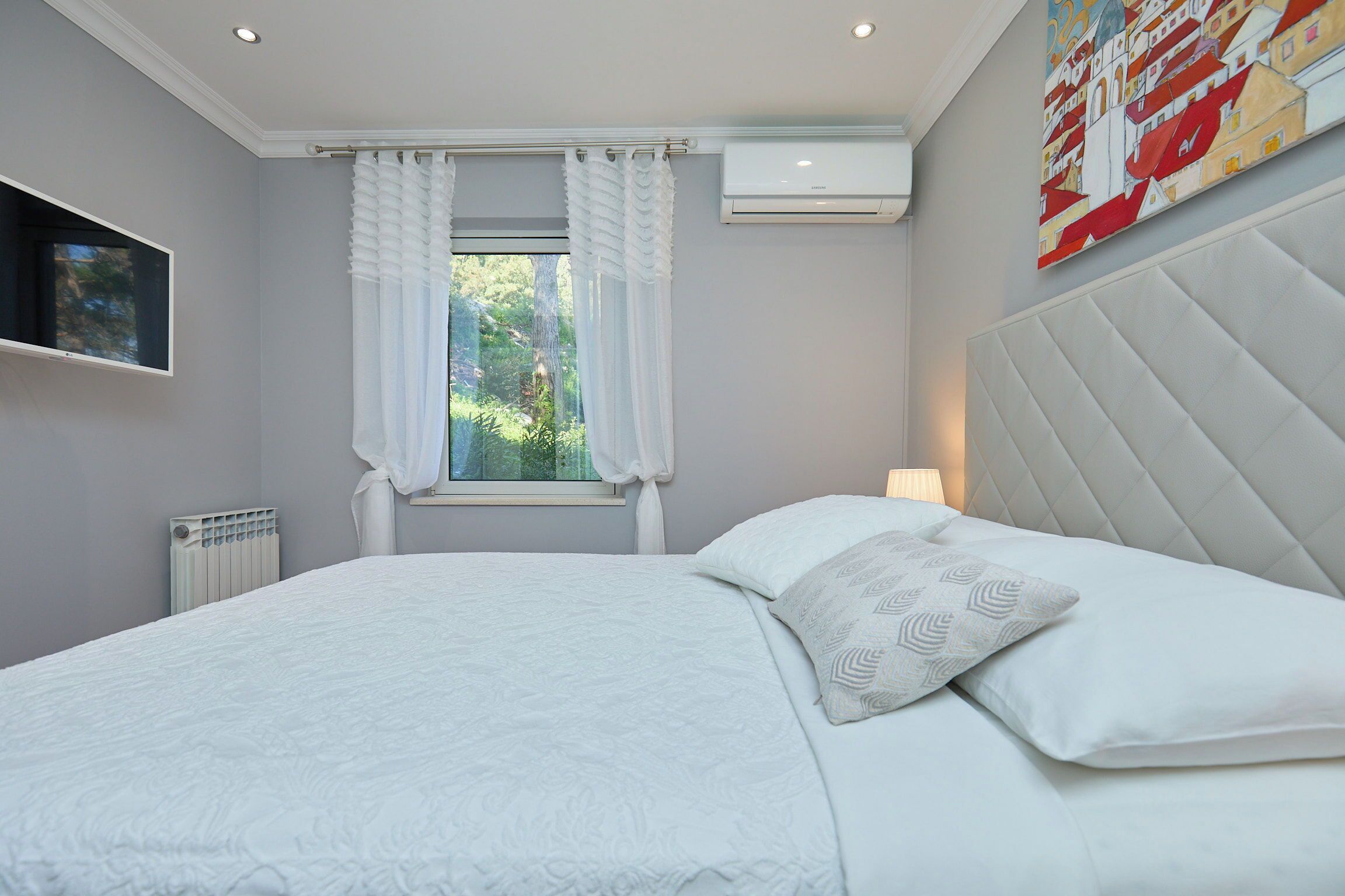 Ein Schlafzimmer mit einem Bett, weißen Vorhängen, einem Fenster, einer Klimaanlage, einem Fernseher und einem Bild an der Wand.