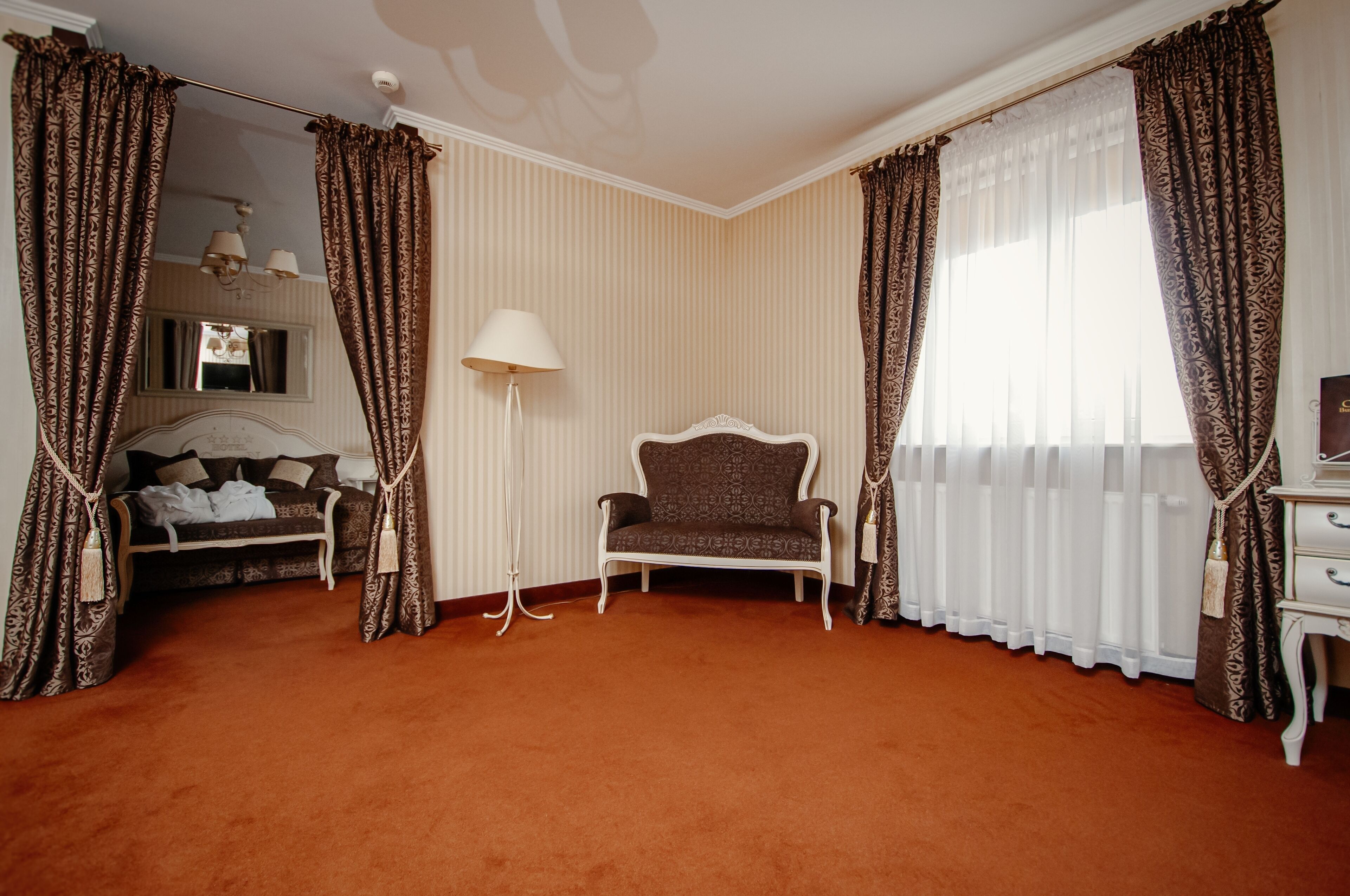 A hotel room with a patterned sofa, a white armchair, a desk, and a large window with curtains.