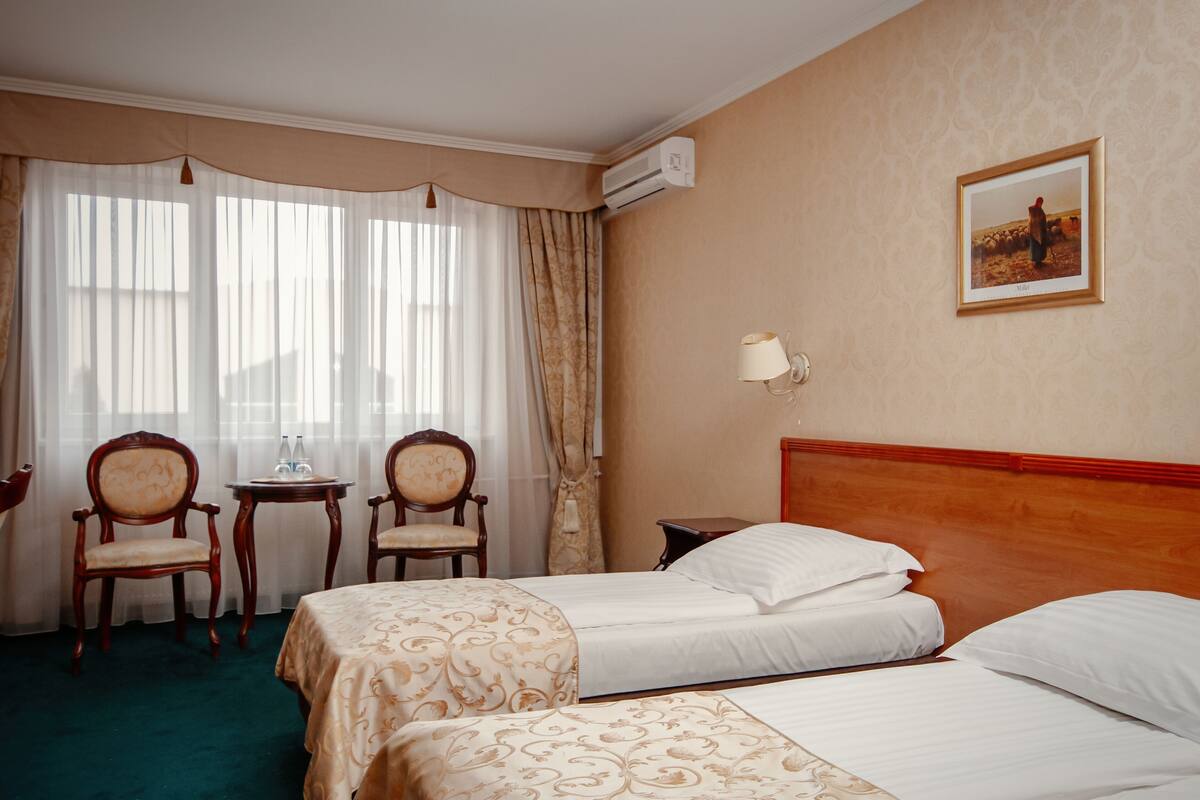 A hotel room with two beds, a small table with chairs, a wall-mounted lamp, and a framed picture on the wall.