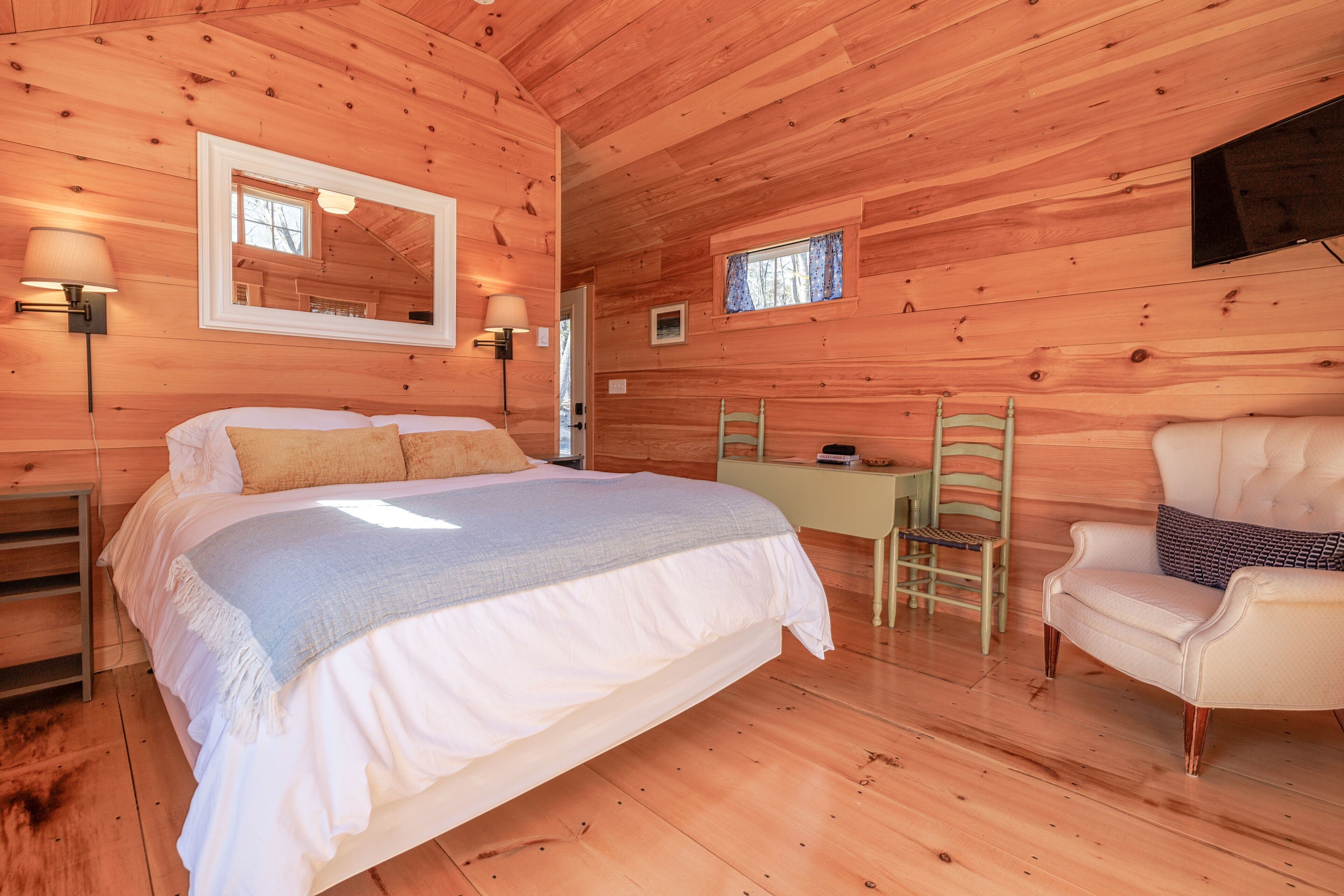 A wooden cabin bedroom with a bed, a chair, a desk, and a TV mounted on the wall.