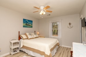 A bedroom with a bed, a ceiling fan, a window, and a small white wicker side table.