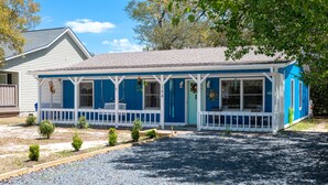 Une maison de plain-pied à l’extérieur bleu et aux ornements blancs, dotée d’un porche avec une balustrade blanche et d’un petit jardin agrémenté d’arbustes.