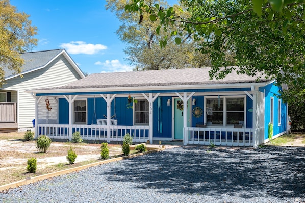 A single-story house with a blue exterior and white trim, featuring a porch with a white railing and a small garden area with shrubs.