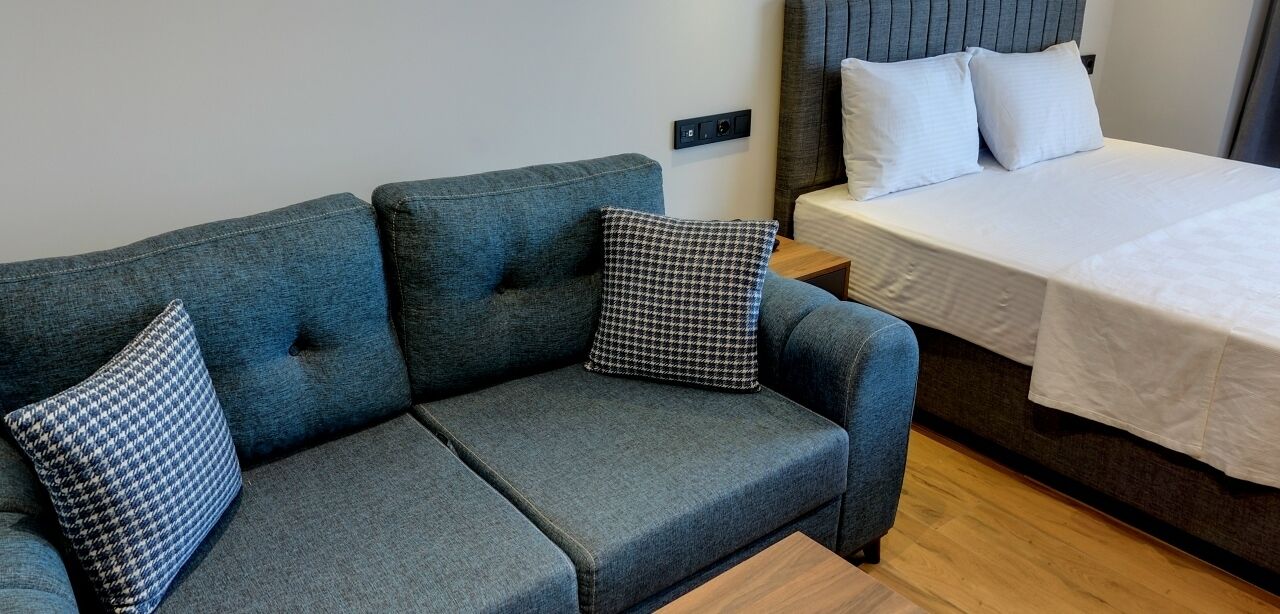 A compact room with a grey sofa, two patterned pillows, a wooden nightstand, and a bed with white bedding.