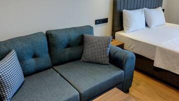 Ein gemütlicher Raum mit einer grauen Couch, zwei gemusterten Kissen, einem hölzernen Nachttisch und einem Bett mit weißer Bettwäsche.