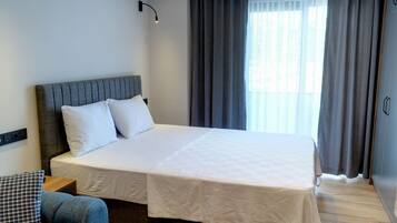 Una cama bien hecha con sábanas blancas, un cabecero tapizado en gris y una mesita de noche de madera con una lámpara.