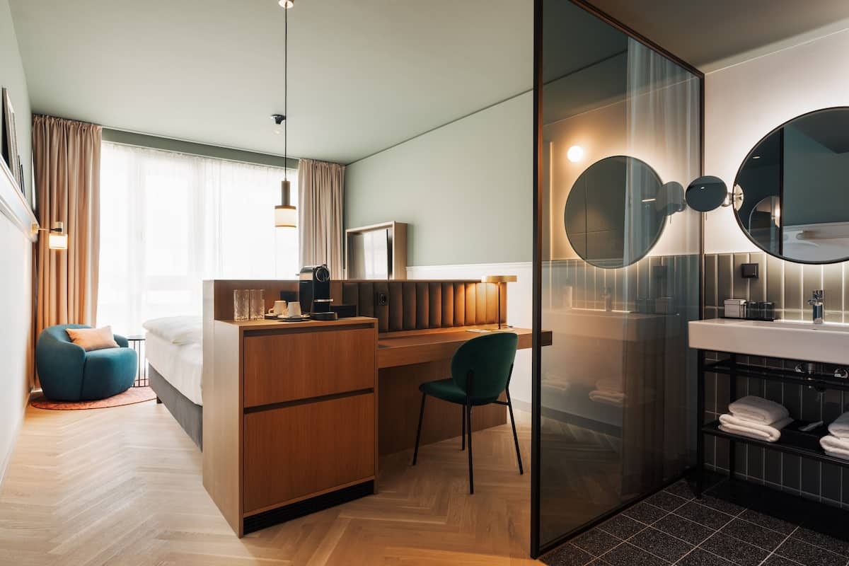 Una habitación de hotel moderna con un escritorio de madera, una silla verde, una cama, un espejo grande y un baño con lavabo y perchero para toallas.