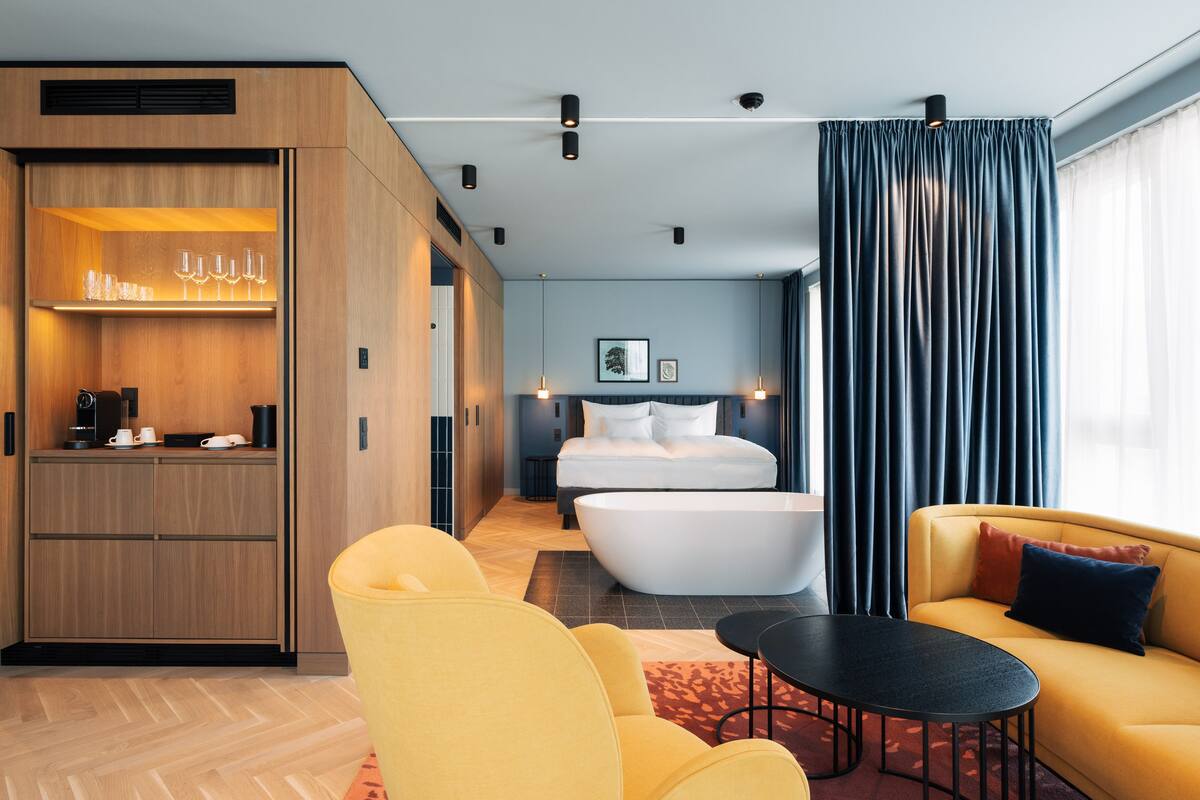 Una habitación de hotel moderna con un armario de madera, una cama grande, un sofá amarillo, una mesa redonda y una bañera.