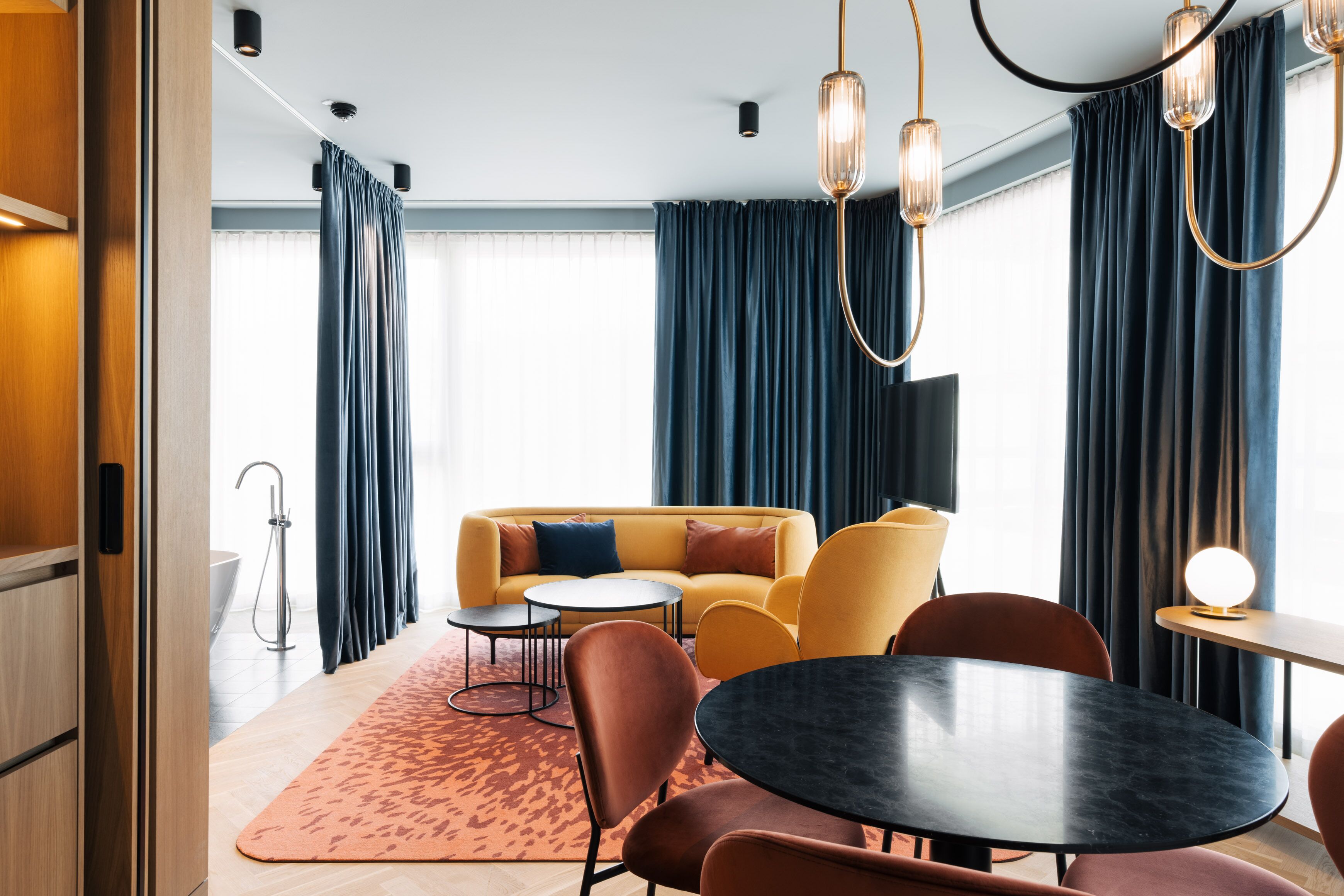 A modern hotel room with a yellow sofa, round table, and chairs, a large window with curtains, and a visible shower area.