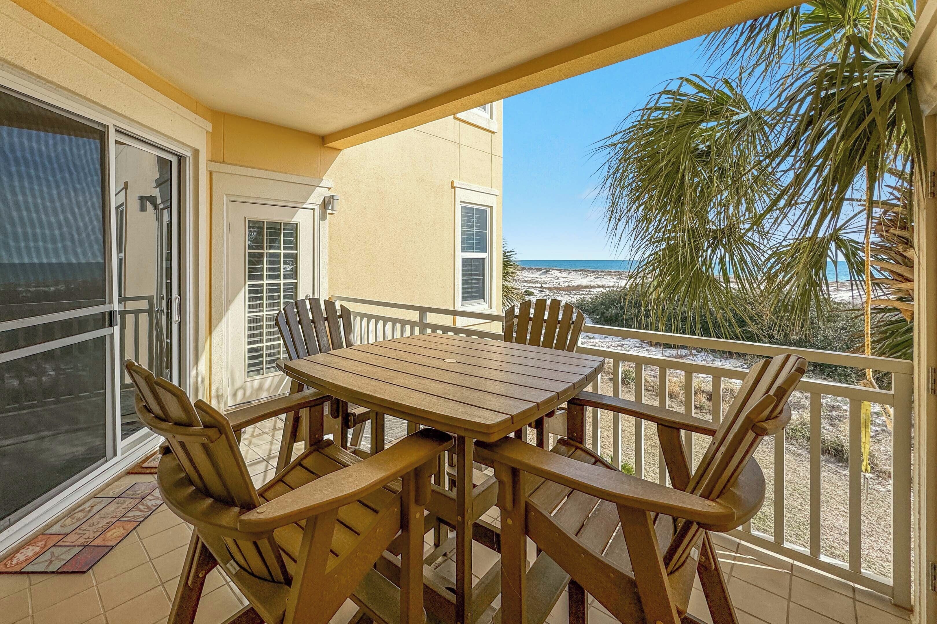 A balcony with a wooden table and chairs, overlooking a beach and palm trees.