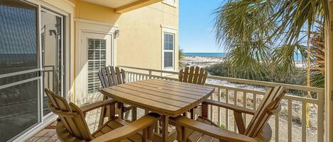 A balcony with a wooden table and chairs, overlooking a beach and palm trees.