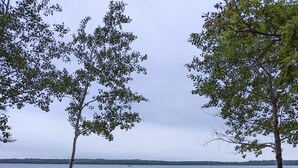 Un área de descanso junto al lago con sillas Adirondack rojas y un hogar para fogata, con vista a un lago tranquilo y la orilla lejana.