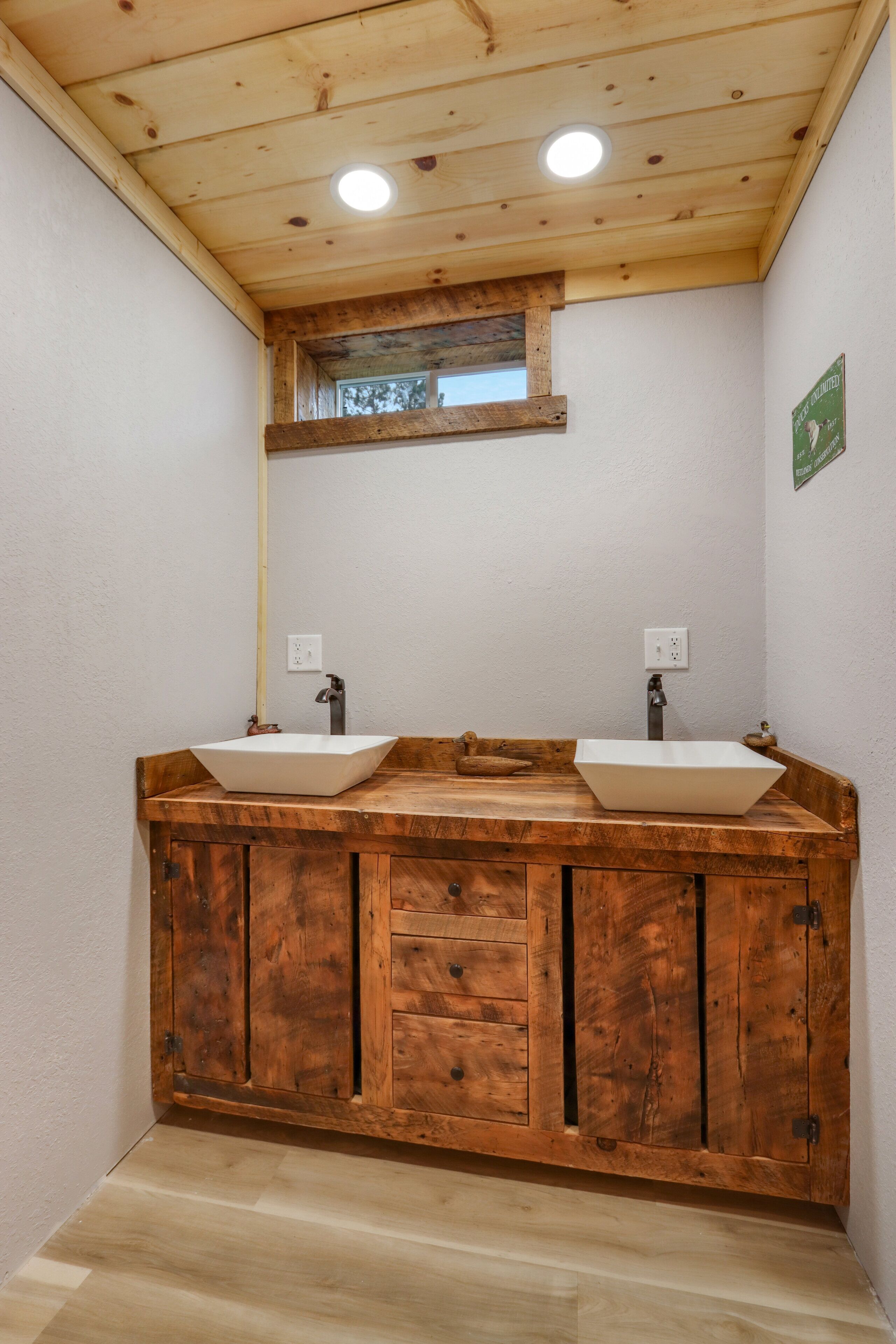 Een badkamer met een houten wastafelmeubel, twee wastafels en een raam boven de spiegel.