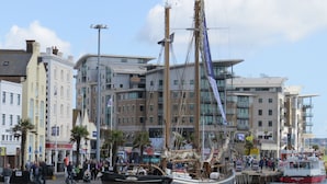 A waterfront area with a large sailing ship docked at a pier, surrounded by modern buildings and a crowd of people. - Andiamo (Poole)