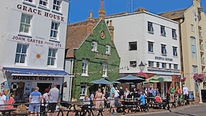 A street scene with buildings labeled 'Grace House' and 'John Carter (Poole) Ltd'. There are outdoor seating areas with tables and umbrellas, and a crowd of people. - Andiamo (Poole)