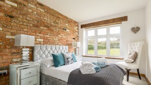 A bedroom with a brick wall, a large window, a bed with a grey patterned bedspread, a white upholstered chair, and a bedside table with a lamp. - Stable Block (Wimborne)